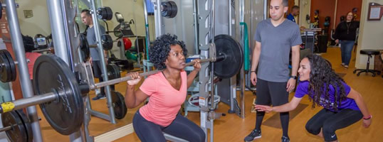 Women doing squats on smith machine with help of a trainer